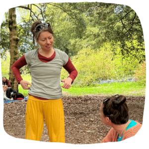 Photo of artist, Holly Thomas, stood in a forest clearing and talking to someone. She is wearing bright yellow trousers with sunglasses on her head.