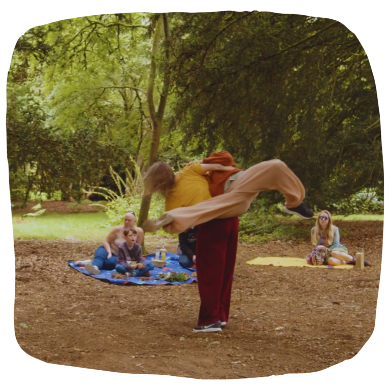 A colour photo of a forest clearing where two dancers are in the middle of a lift. One dancers appears to be leaping through the air, supported by her partner's back. Groups of a people are watching, sat on coloured rugs and surrounded by tall green trees.