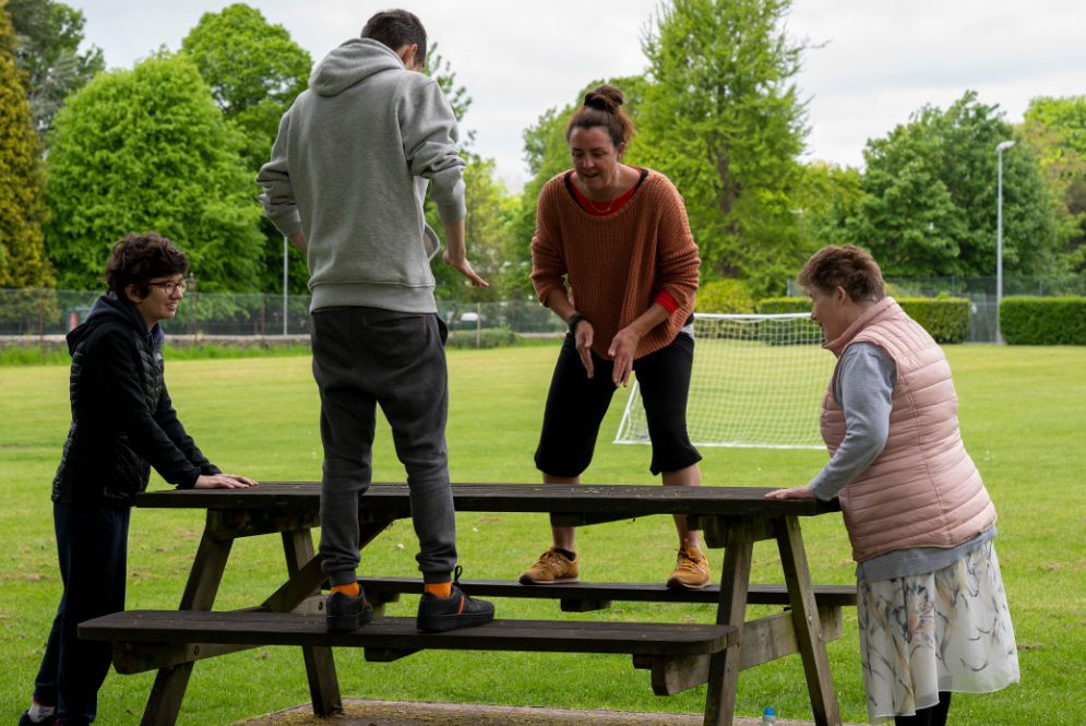 A colour photo of 3 participants with facilitator Louise around a picnic bench in a park. Two are stood on seats, two stood at the end. They are clapping their hands in the air and on the bench.