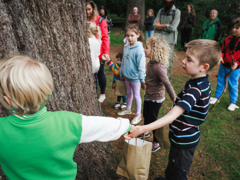 A colour photo of a group of children holding hands to make a circle around a tree.